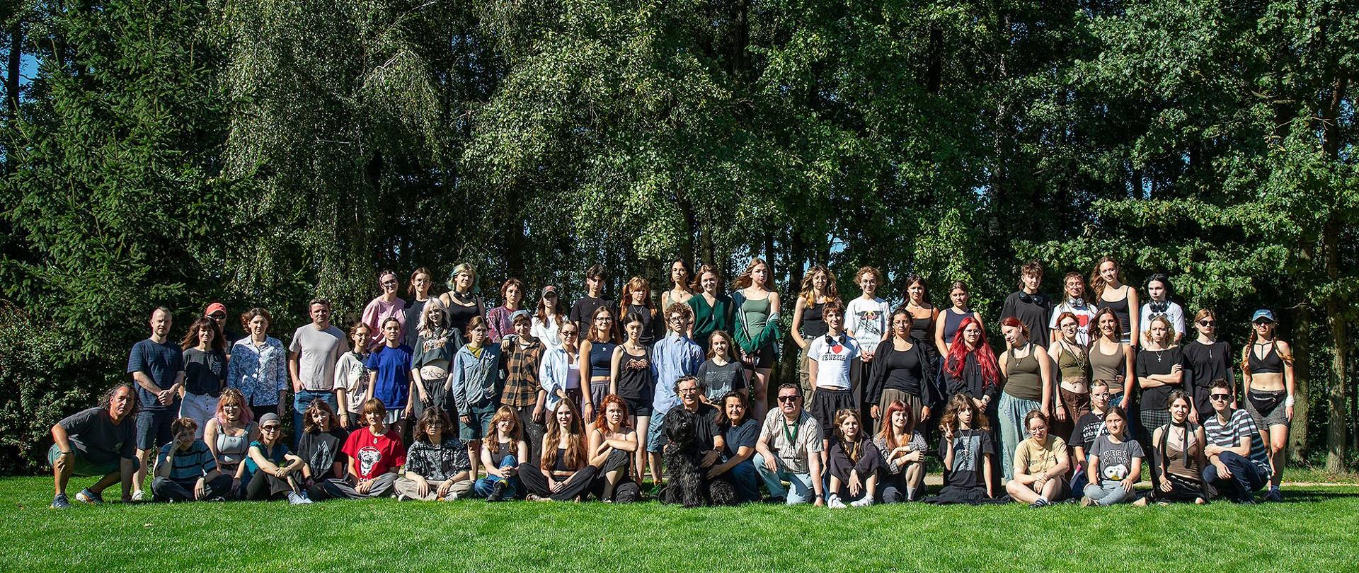 Duże, grupowe zdjęcie około 60-70 osób, głównie młodych dorosłych, pozujących na zielonym trawniku na tle gęstych drzew i krzewów w słoneczny dzień. Większość osób siedzi lub kuca w pierwszym rzędzie, a pozostałe stoją w dwóch rzędach za nimi. Wszyscy ubrani są w swobodne, letnie stroje.