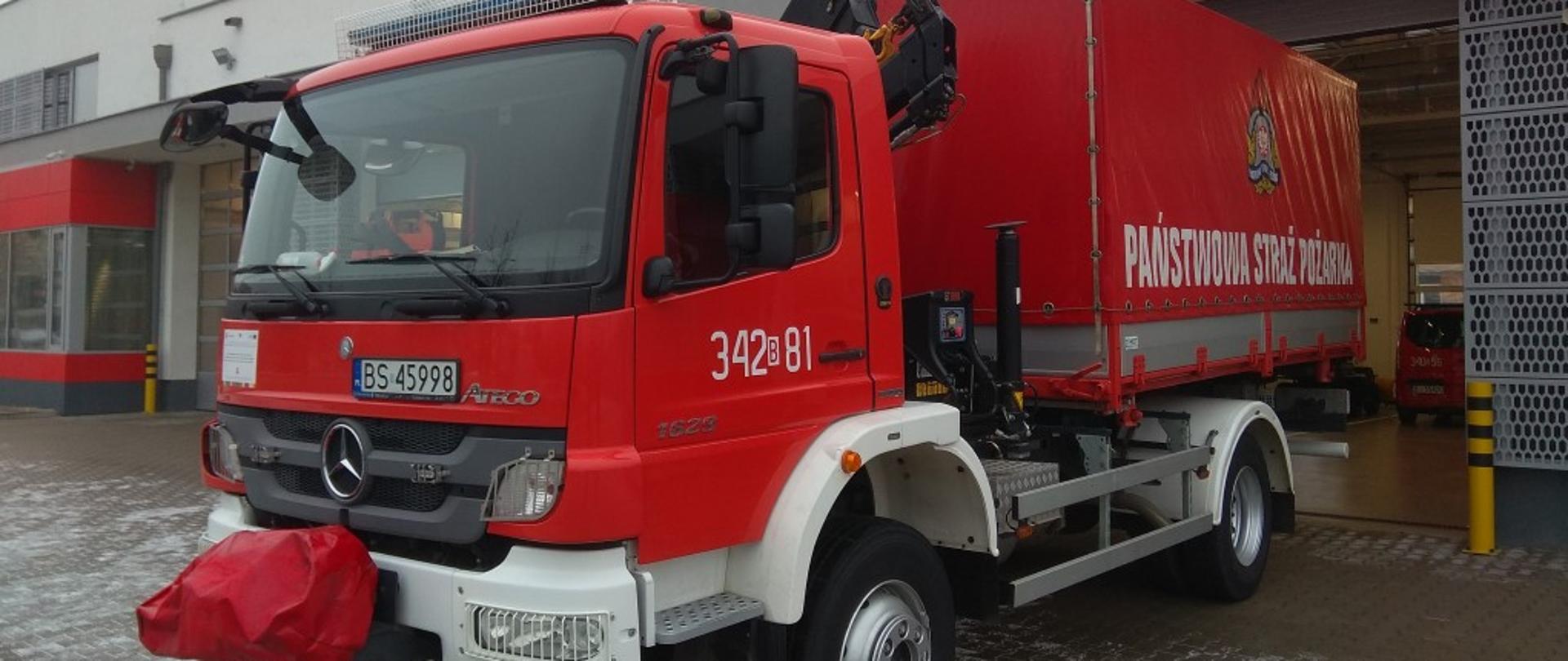 Na pierwszym planie samochód typu ciężkiego kwatermistrzowski do przewozu sprzętu pożarniczego. Kolor pojazdu czerwony z białym zderzakiem. W tle garaże Jednostki Ratowniczo-Gaśniczej nr 2