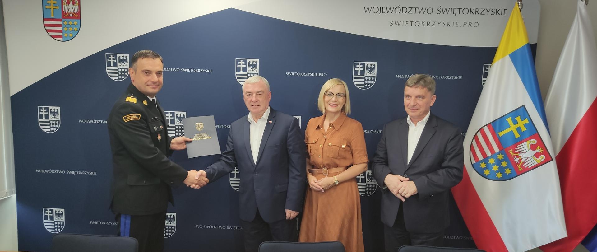 Na zdjęciu widzimy 4 osoby. 3 mężczyzn i kobietę. Od lewej: dwóch mężczyzn wymienia uścisk dłoni i teczkę. Jeden z nich jest w mundurze generała PSP drugi w garniturze ciemnym. Kobieta w brązowej sukience, a ostatni stoi mężczyzna w garniturze ciemnym. 