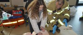 Ćwiczenia w multibibliotece. Korytarz na pierwszej kondygnacji. Jedna z pracownic przy instruktażu strażaka prowadzi ucisk klatki piersiowej manekina. W tle sprzęt ratownictwa medycznego.