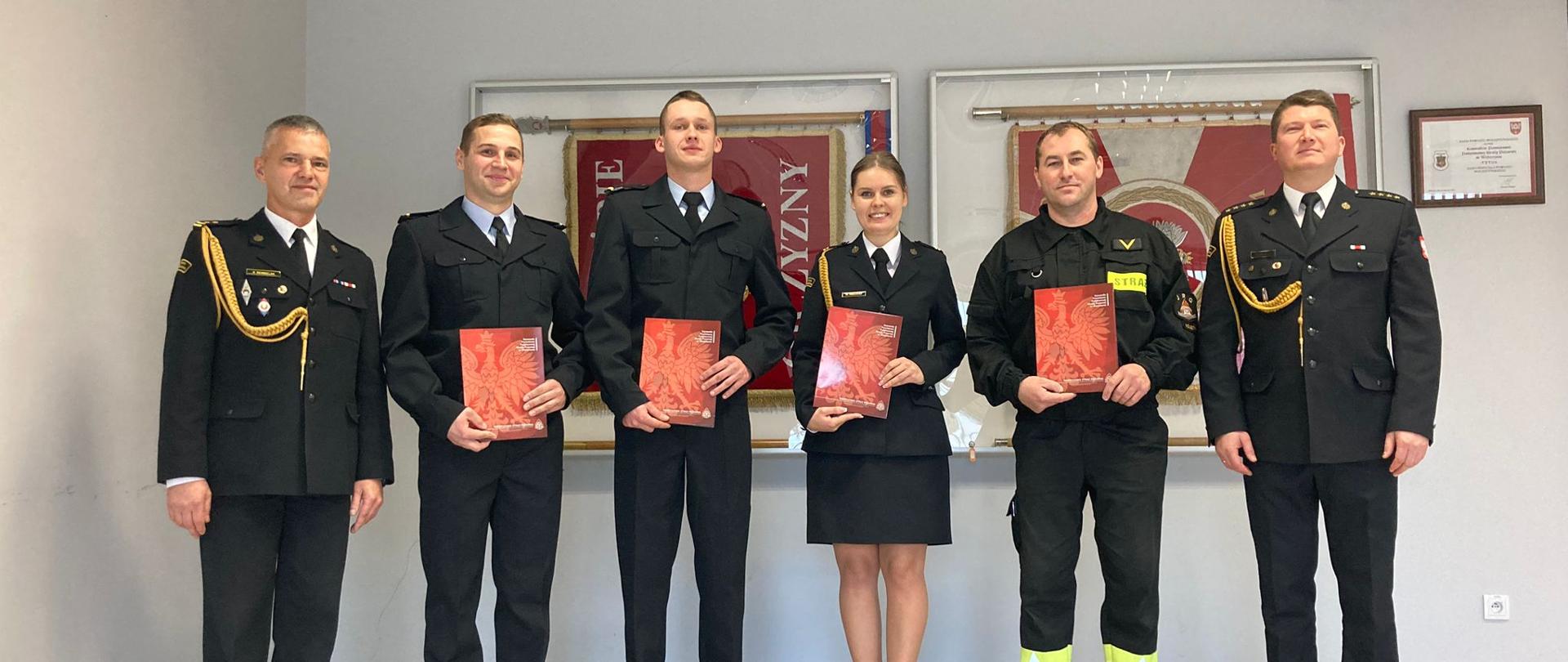 Zdjęcie przedstawia Komendanta Powiatowego PSP w Wolsztynie wraz z zastępcą oraz czterema awansowanymi strażakami. W tle gabloty ze strażackimi sztandarami.