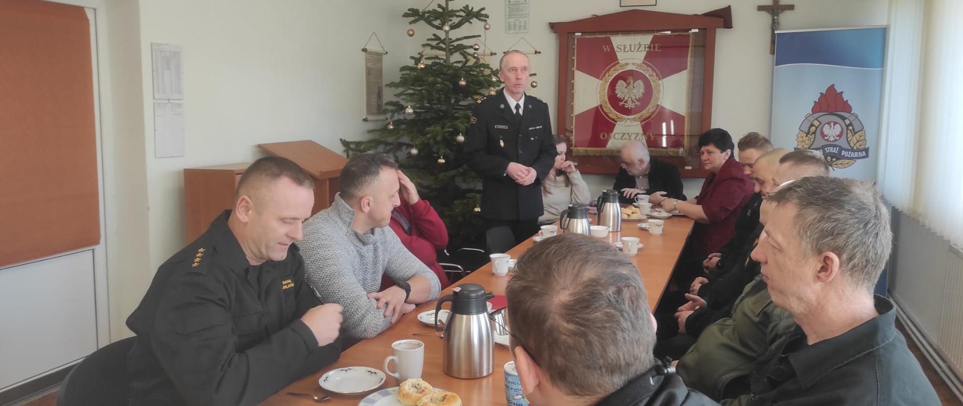 W centrum zdjęcia znajduje sie stół przy którym siedzą strażacy oraz przedstawiciele z gmin. W tle widać choinkę, sztandar KP PSP Świdwin oraz Godło i Krzyż.