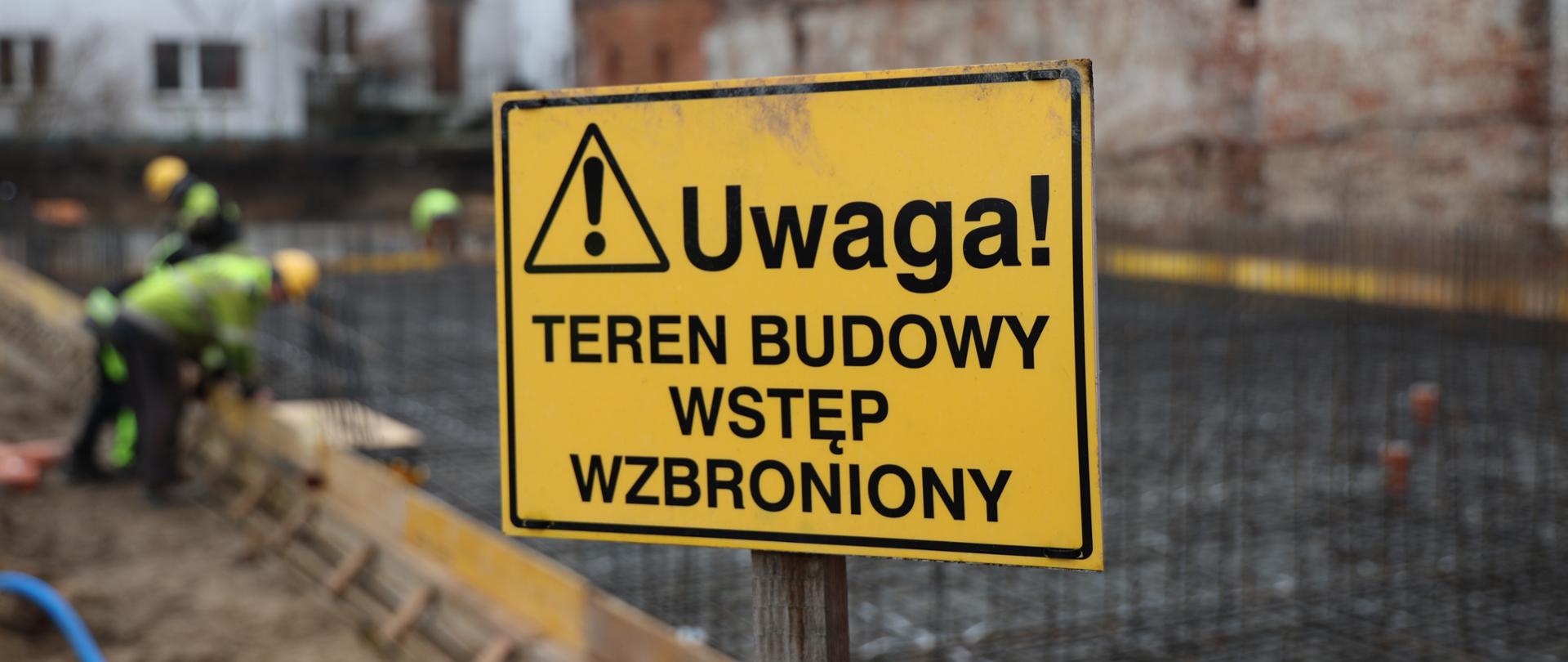 Tablica w kolorze żółtym z czarnym napisem - Teren budowy wstęp wzbroniony. Za nią rozmazany obraz starego muru i pracowników budowy,
