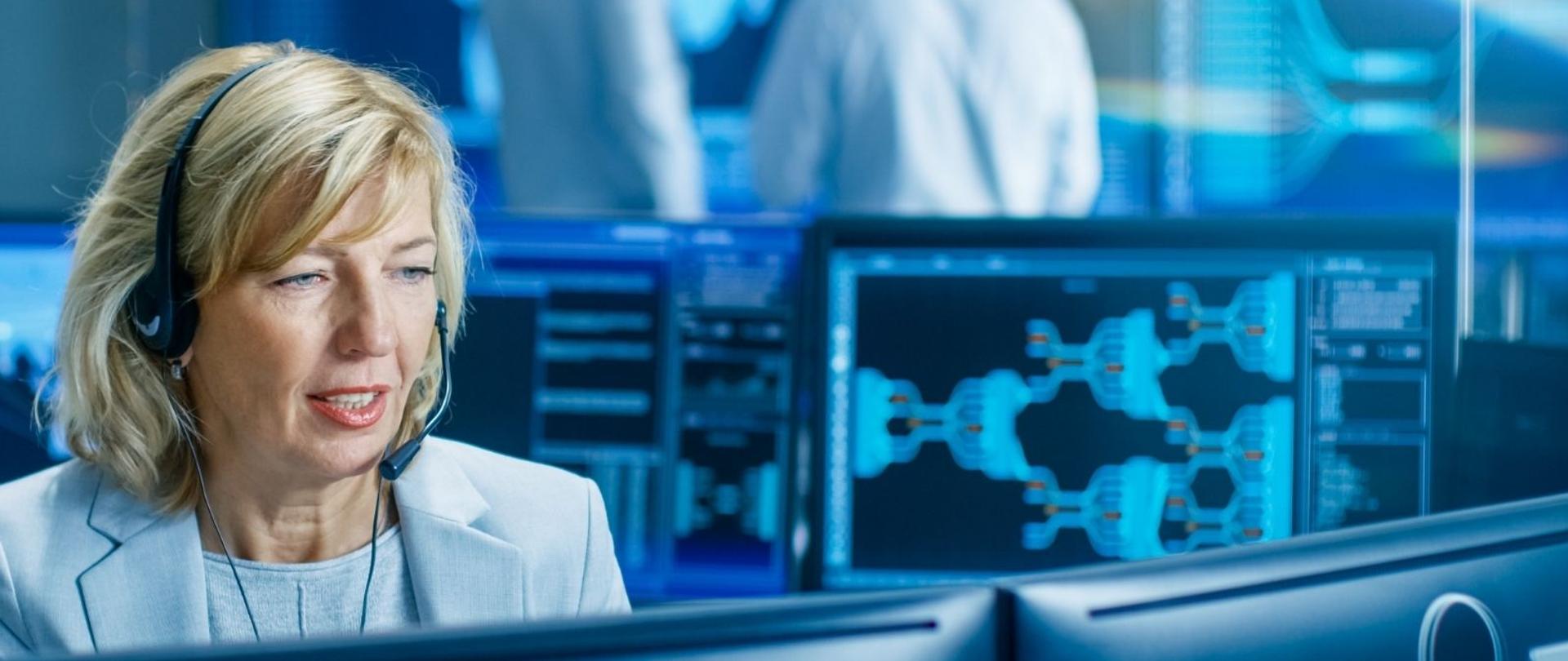 Na zdjęciu kobieta ze słuchawką na uchu i mikrofonem, przyjmująca zgłoszenie. 