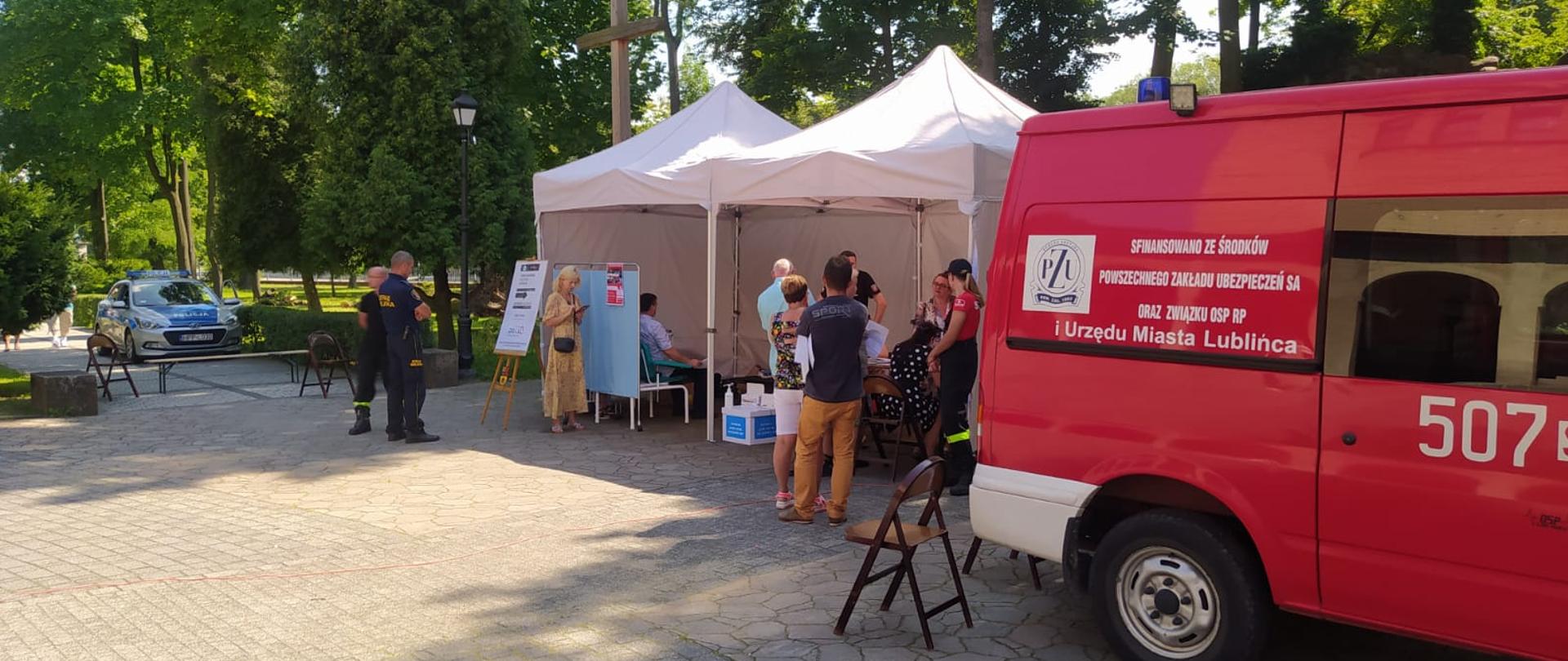 Zdjęcie tymczasowego punktu szczepień przy parafii kościelnej. Po prawej stronie samochód pożarniczy. Za samochodem białe namioty oraz kilkoro ludzi do szczepień przeciw koronawirusowi. W tle wysokie drzewa oraz samochód policyjny.