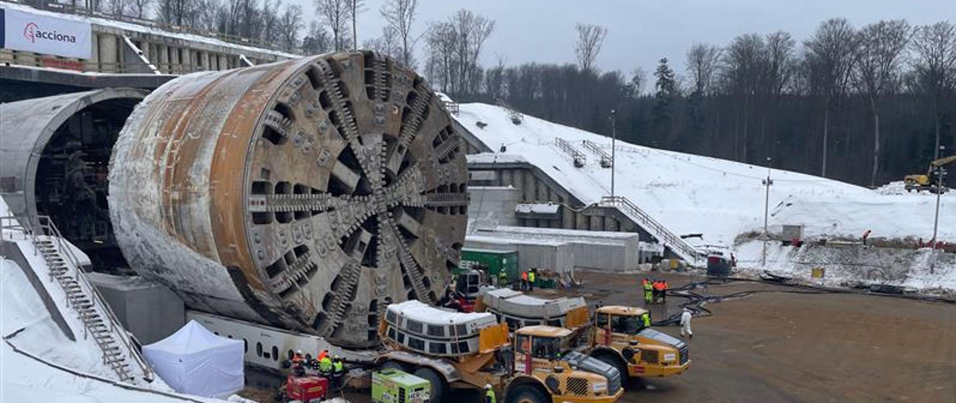 Duża tarcza TBM do drążenia tuneli ustawiona na placu budowy przy wylocie tunelu. Wokół znajdują się maszyny budowlane, pracownicy w odzieży ochronnej oraz zaśnieżony teren.
