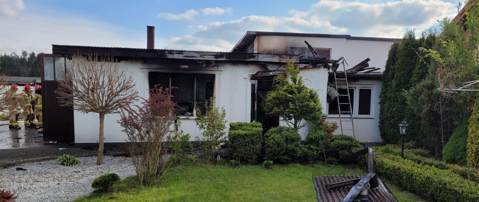 Zdjęcie przedstawia budynek warsztatowy po pożarze. W otwartych wrotach garażowych stoi grupa strażaków.