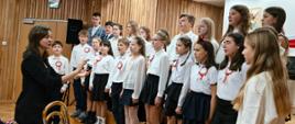 Duży chór dzieci i młodzieży, ubranych na biało-czarno z biało-czerwonymi kotylionami, śpiewa, stojąc w rzędach. Przed nimi stoi dyrygentka w czarnym żakiecie, kierując występem.