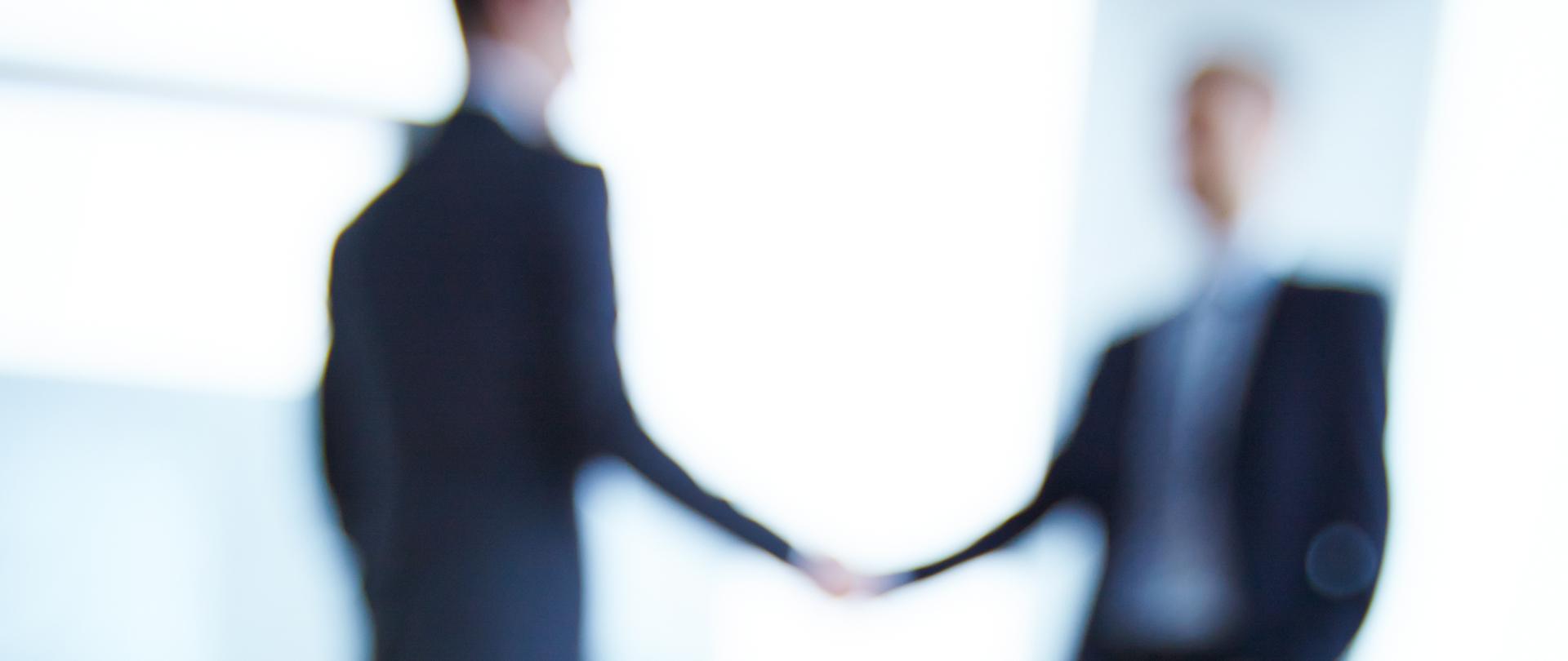 Business document and pen on desk with two employees handshaking on background