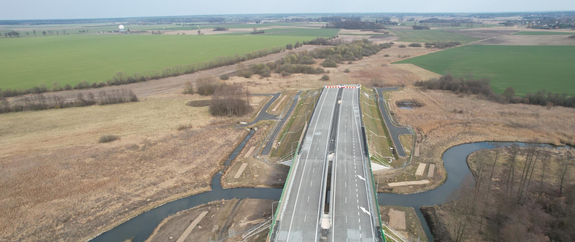 Zdjęcie przedstawia widok z lotu ptaka na nowo wybudowany odcinek autostrady. Autostrada ma kilka pasów ruchu i jest otoczony polami oraz fragmentami drzew. Krajobraz jest głównie płaski z zielonymi i brązowymi plamami, wskazującymi na różne rodzaje roślinności lub użytkowania terenu.