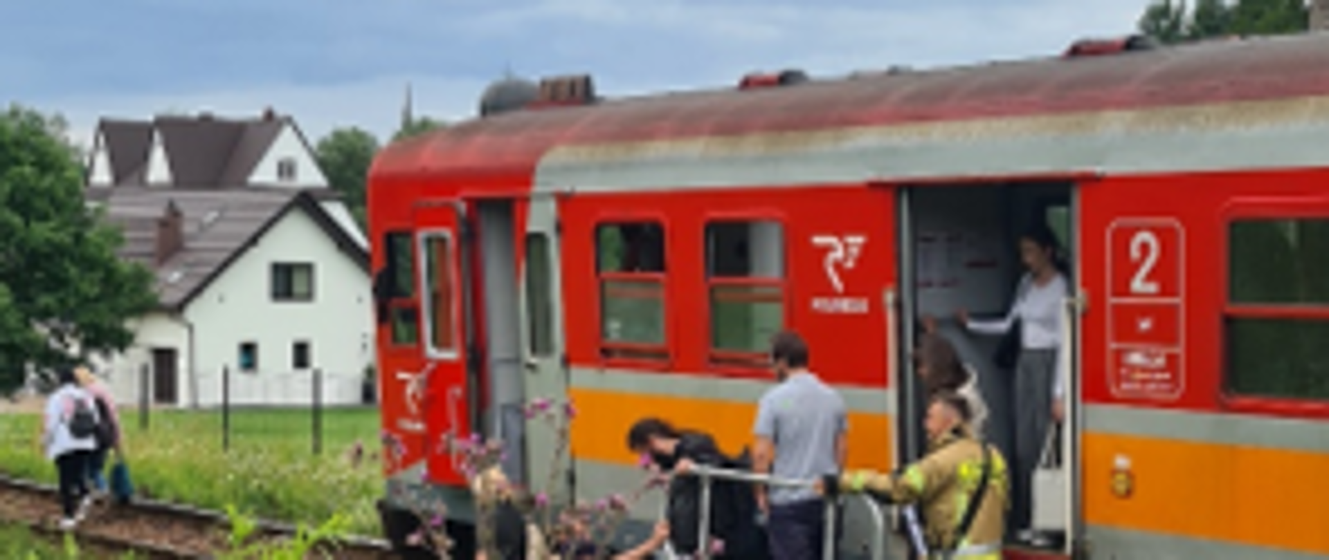 Zdjęcie przedstawia skład kolejowy który uległ awarii. Na pierwszym planie za zaroślami widać pasażerów którzy przy pomocy strażaków po podeście ratowniczym opuszczają pociąg i z bagażami wzdłuż linii kolejowej udają się w miejsce zbiórki.