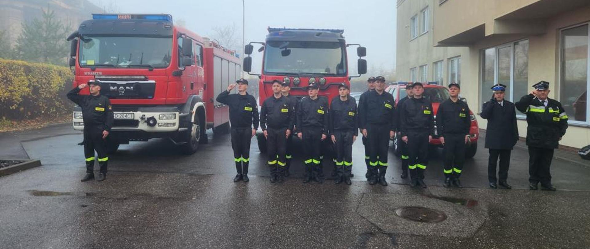 Ustawieni w dwuszeregu strażacy przed samochodami pożarniczymi 