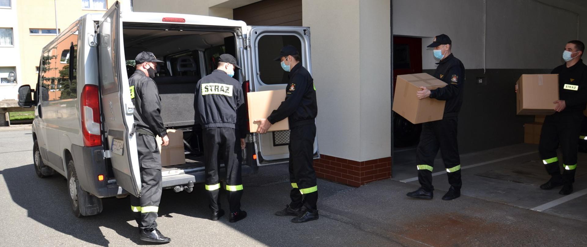 Na zdjęciu 5 strażaków w ubraniach koszarowych przenosi pudełka z maseczkami jednorazowymi do samochodu dostawczego.