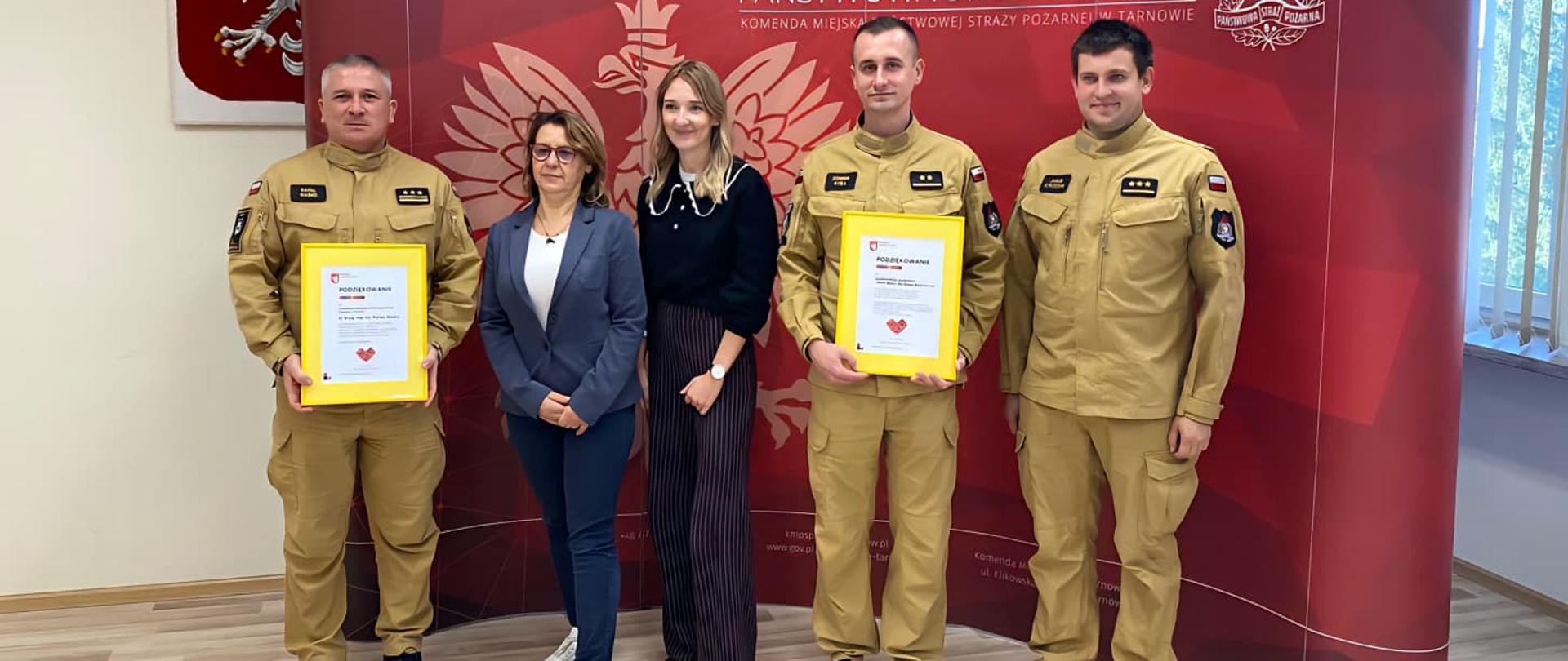 W rzędzie stoją strażacy ubrani w piaskowe mundury oraz przedstawiciele fundacji, strażacy trzymają w rękach dyplomy. Z tyłu ścianka reprezentacyjna PSP oraz godło Polski nad którym wisi krzyż