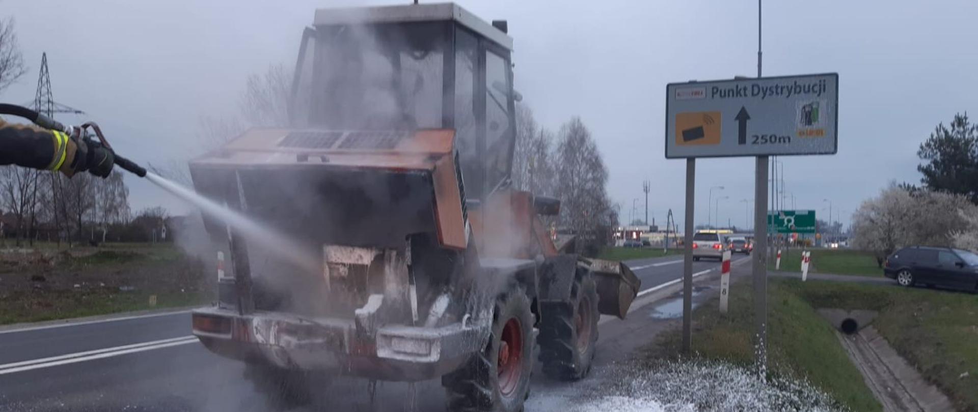 Przy ulicy serbskiej w Żarach trwa akcja gaśnicza palącej się ładowarki. Strażacy gaszą pianą palący się tył pojazdu. Na poboczu widać sześć zużytych gaśnic proszkowych.