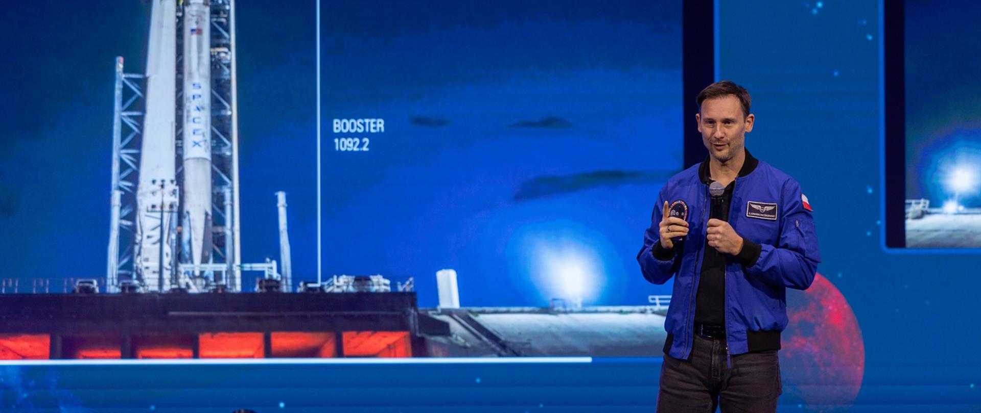 Sławosz Uznański-Wiśniewski w niebieskiej kurtce mówi do trzymanego w ręku mikrofonu, stoi na tle niebieskiej ścianki na której z lewej widać zdjęcie rakiety na stanowisku startowym.