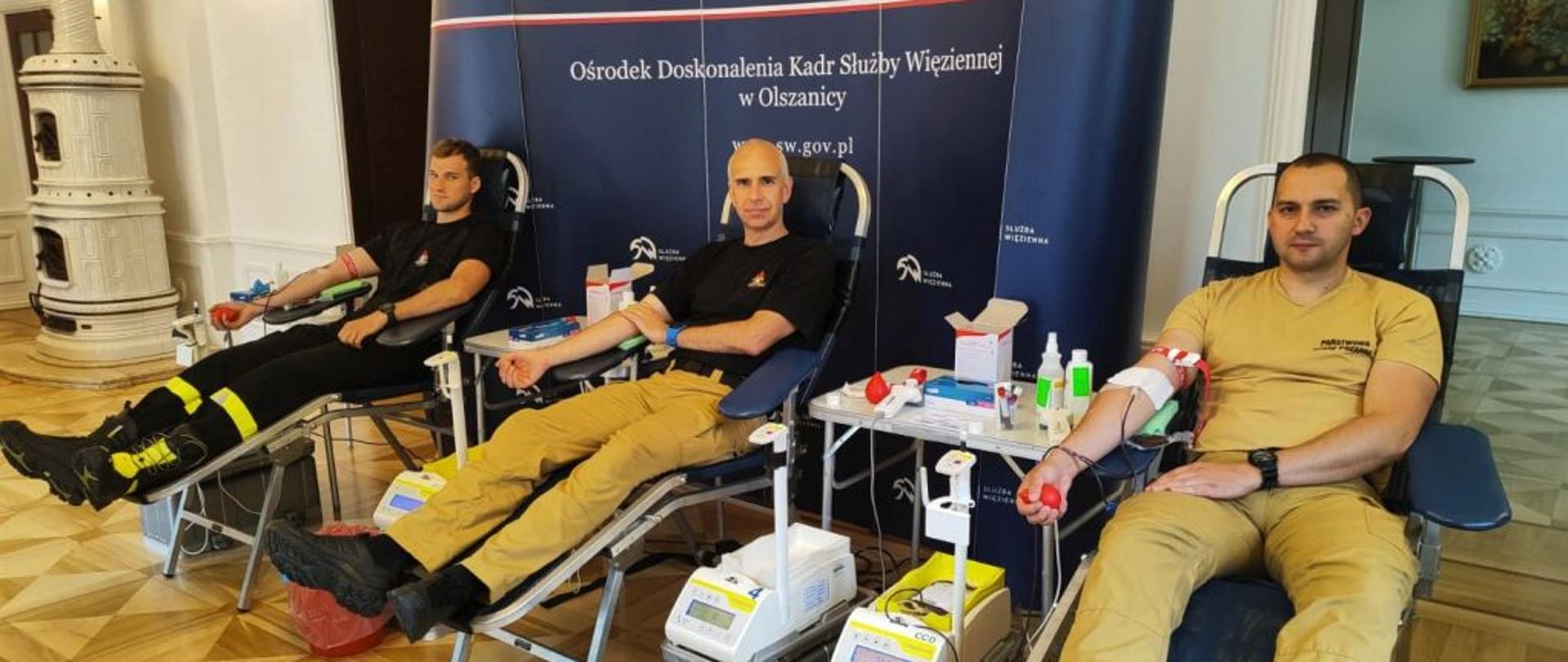 Na zdjęciu widać zastępce komendanta powiatowego PSP w Ustrzykach Dolnych, stażystę oraz kadrowca z tej samej komendy którzy siedzą na krzesłach do oddawania krwi. Są oni ubrani w piaskowe mundury z wyjątkiem czarnej koszulki u kadrowca i ubrania koszarowego u stażysty. Na rękach zaciśnięte mają opaski a w dłoniach piłeczkę do ściskania. obok stoi jedno krzesło które jest wolne. Za nimi stoi baner Straży Więziennej 