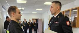 Na zdjęciu w mundurze galowym po prawej komendant powiatowy wrączający akt ślubowania strażakowi składającemu ślubowanie. Strażak jest ubrany w ubranie czarne koszarowe. Uroczystość ślubowania ma miejsce w sali konferencyjnej.