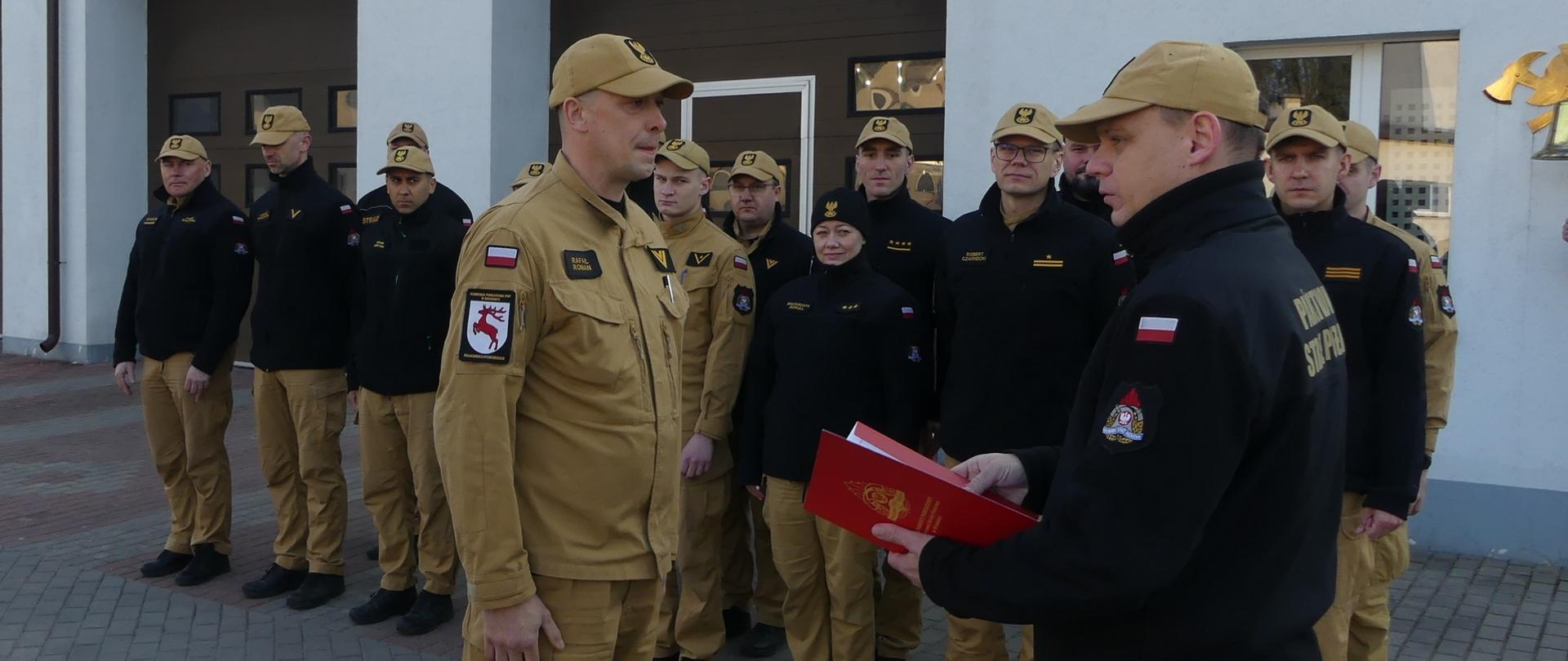 Zdjęcie przedstawia starszego ogniomistrza Rafała Romana podczas wręczenia mianowania na stanowisko Starszy operator sprzętu. Mianowanie wręcza Komendant Powiatowy PSP w Brodnicy. W tle w dwuszeregu stoją funkcjonariusze Komendy.