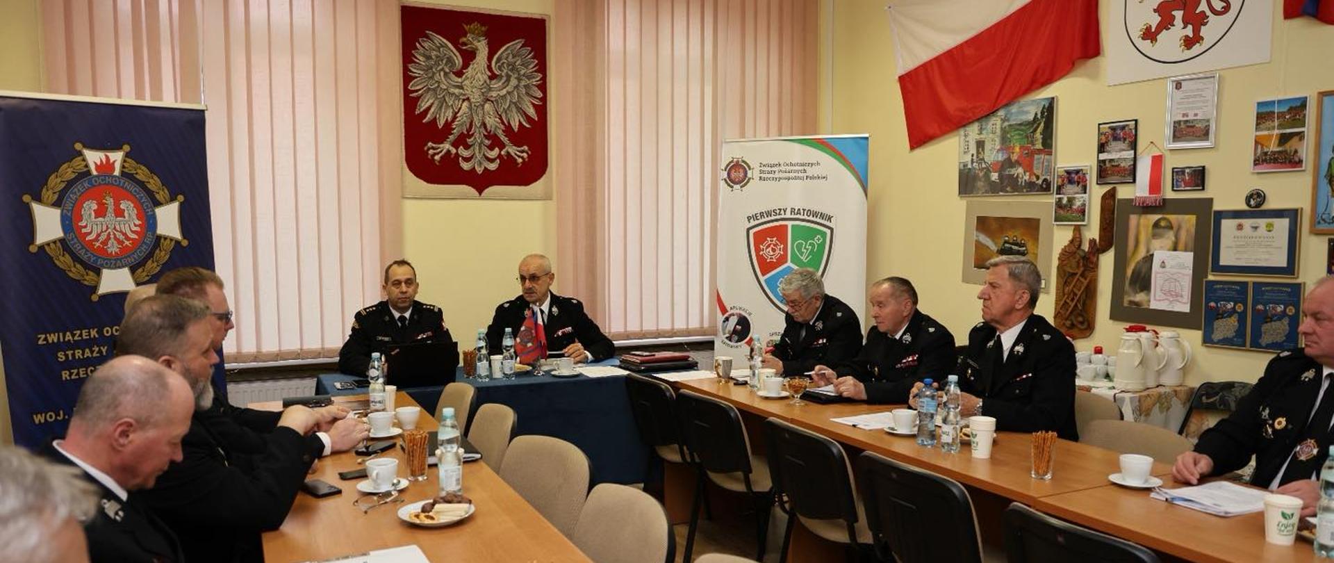 Na zdjęciu widać spotkanie w sali konferencyjnej. Przy długich stołach siedzą mężczyźni w mundurach strażackich, rozmawiają i notują. Na ścianach wiszą godło Polski, flaga oraz plakaty i zdjęcia. Na stołach stoją kubki, woda i dokumenty. Atmosfera jest oficjalna.