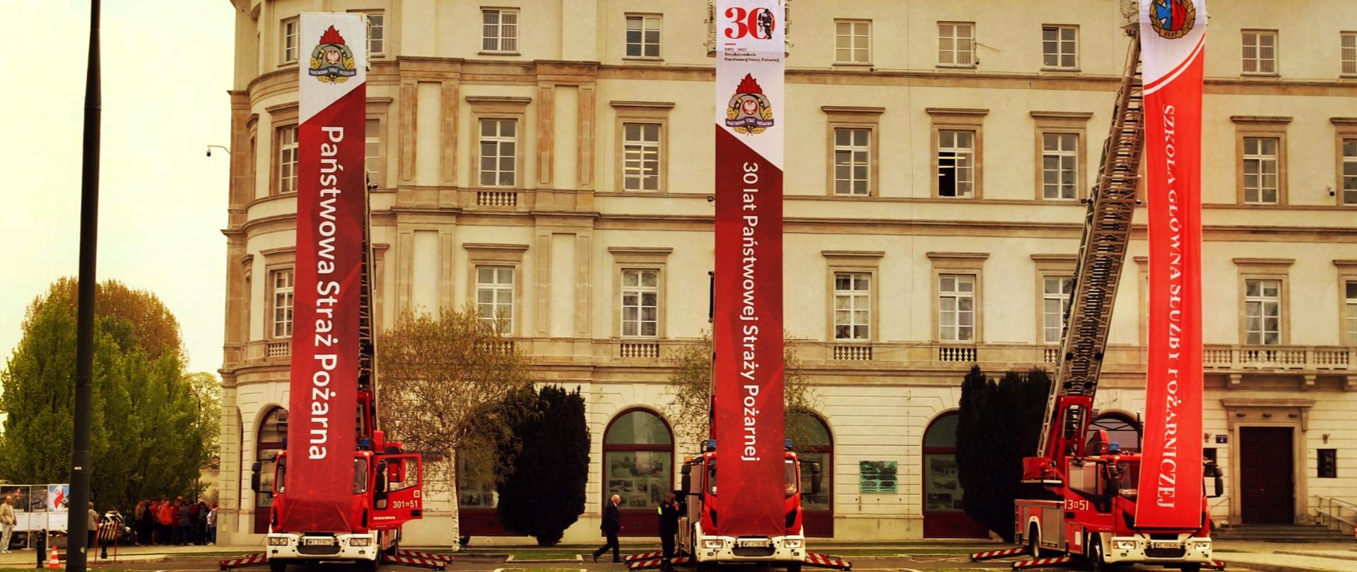 Na zdjęciu trzy samochody gaśnicze drabiny stojące blisko siebie w równych odstępach. Każdy ma rozwiniętą do góry drabinę z której zwisają banery reklamowe