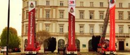 Na zdjęciu trzy samochody gaśnicze drabiny stojące blisko siebie w równych odstępach. Każdy ma rozwiniętą do góry drabinę z której zwisają banery reklamowe