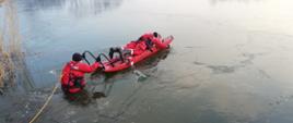 Zdjęcie przedstawia dwóch strażaków w wodzie. Jeden ze strażaków leży na desce wypornościowej przeznaczonej do działań na wodzie i lodzie koloru czerwonego, drugi strażak stoi w wodzie po tym jak lód się załamał i popycha deskę do przodu. 