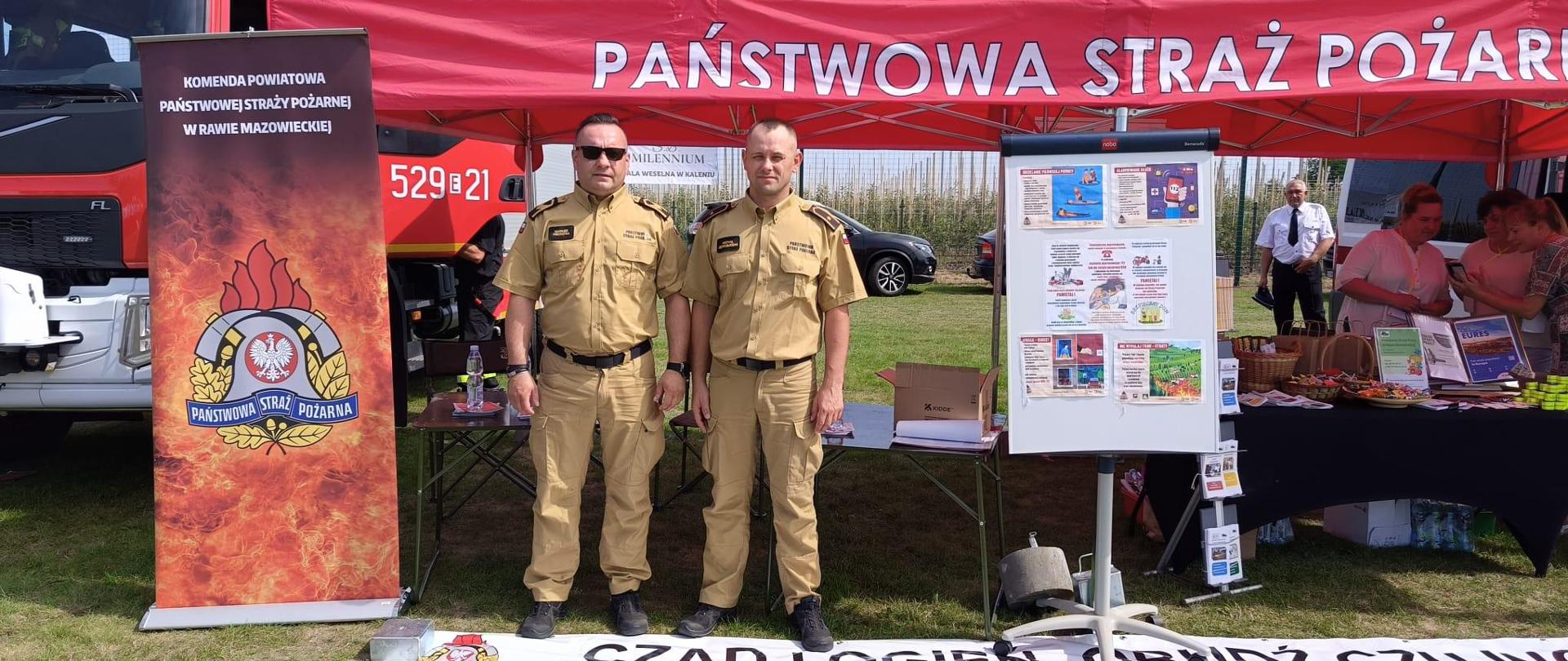 Dwóch strażaków ubranych w umundurowanie służbowe, w kolorze piaskowym, stoi przed stoiskiem Państwowej Straży Pożarnej, które zostało rozstawione, aby promować akcję „Czad i Ogień. Obudź Czujność”. Za nimi znajdują się samochody strażackie.