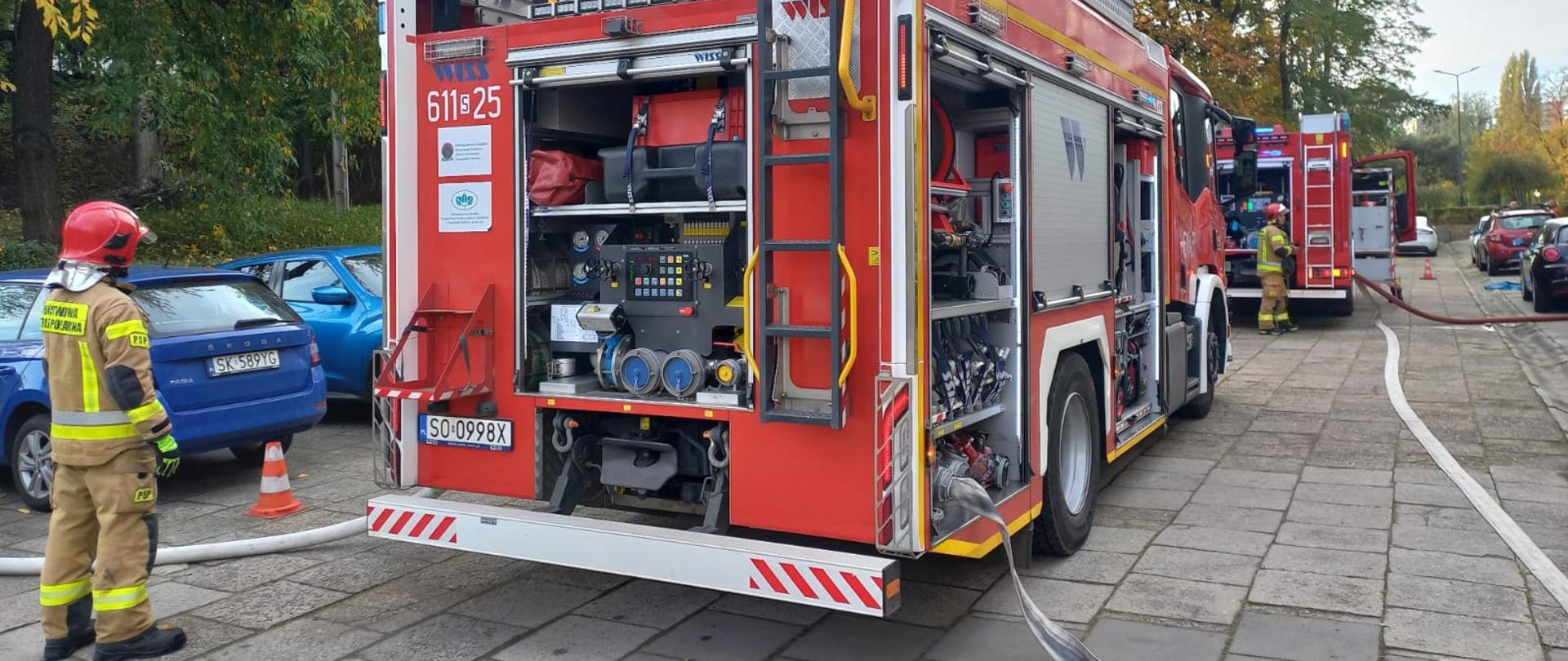Na zdjęciu widać strażaków oraz wozy strażackie Państwowej Straży Pożarnej ustawione wzdłuż ulicy podczas ćwiczeń. Jeden ze strażaków stoi obok pojazdu z otwartymi przedziałami sprzętowymi, z których wyciągnięte są węże i wyposażenie ratownicze. W tle widoczne są drzewa w jesiennych barwach oraz zaparkowane samochody