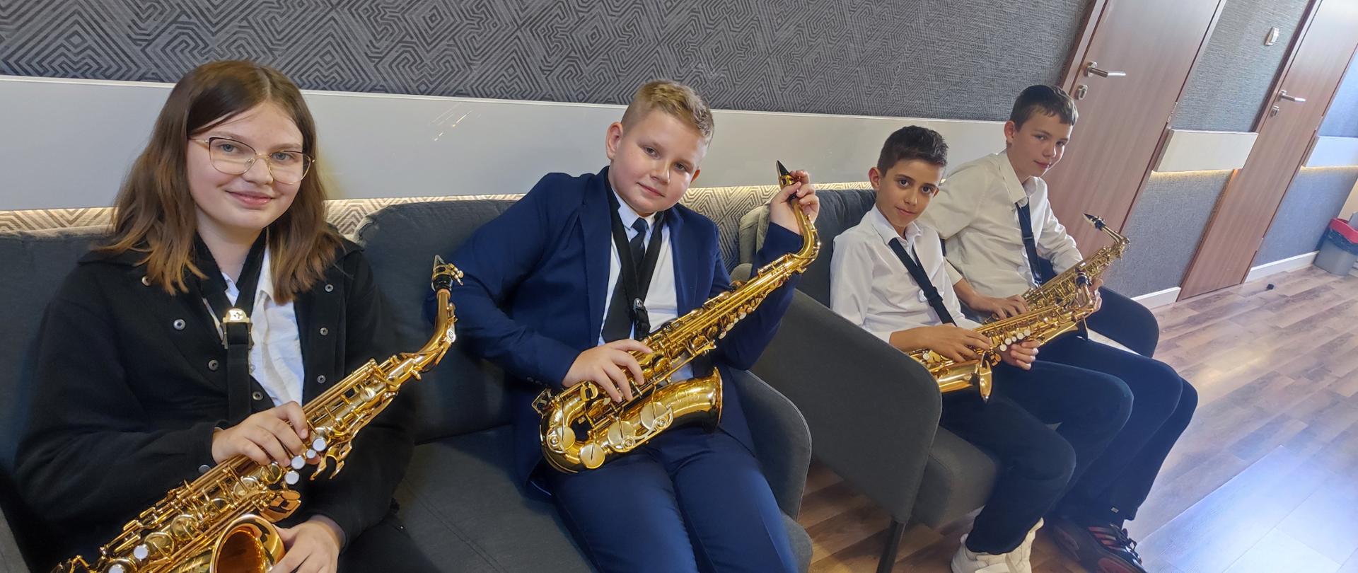 Uczniowie w strojach galowych siedzą na fotelach, każdy trzyma saksofon, uśmiechają się