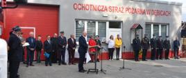 Zdjęcie przedstawia zaproszonych gości na uroczystość ostawionych obok siebie wzdłuż budynku remizy.