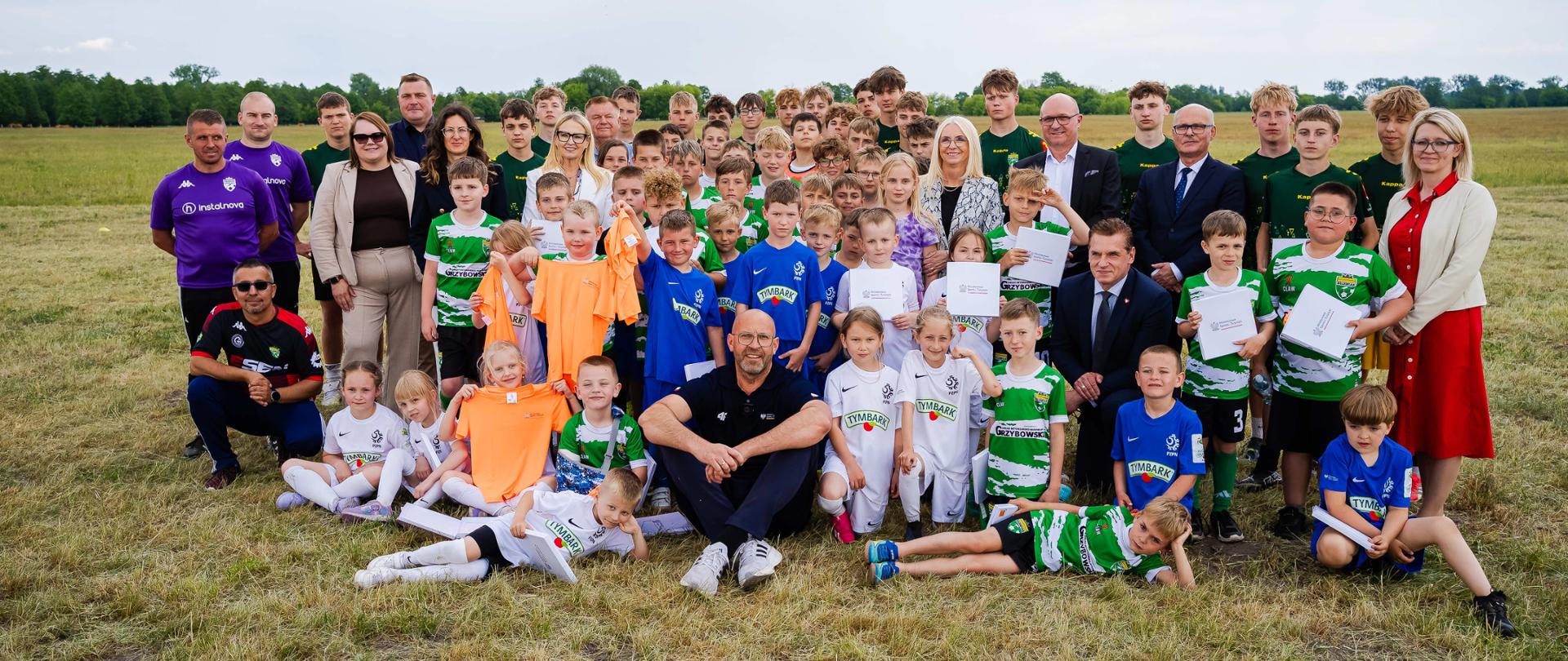 Zbiorowe zdjęcie ministra sportu i turystyki Jakuba Rutnickiego z młodymi zawodnikami i uczestnikami konferencji w Kruszynie. Zdjęcie zrobione na polu. W tle widać drzewa.