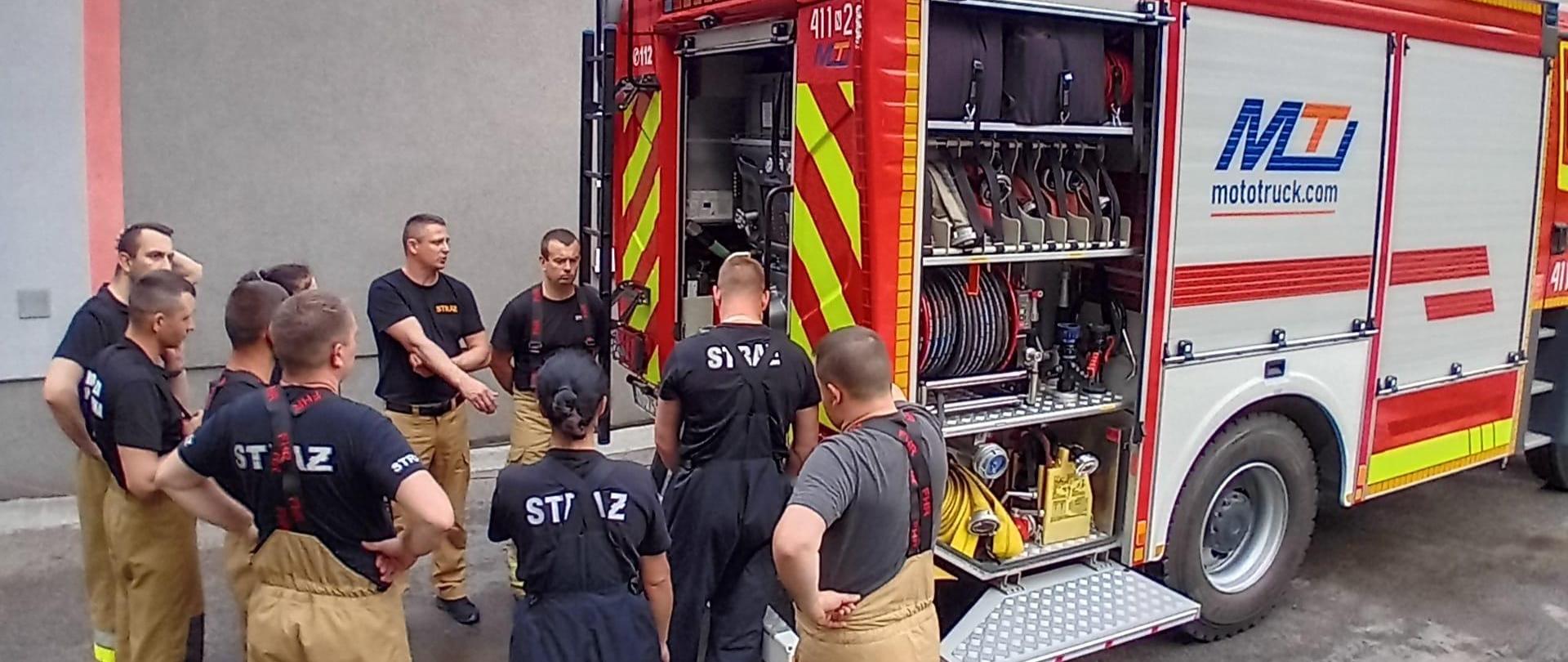 Strażacy przy samochodzie strażackim, trwa szkolenie, omawianie sprzętu.