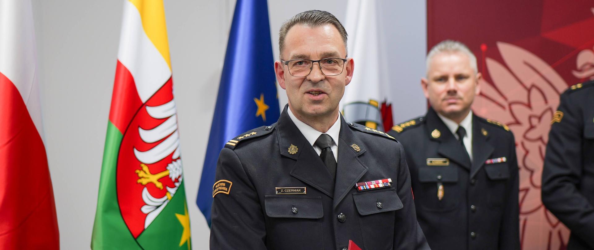 Portretowe zdjęcie starszego brygadiera Zbigniewa Czerniaka w mundurze wyjściowym z licznymi baretkami odznaczeń na piersi. Oficer trzyma czerwoną teczkę i patrzy prosto w kamerę, lekko się uśmiechając. W tle widoczne są flagi: Polski, województwa lubuskiego oraz Unii Europejskiej.