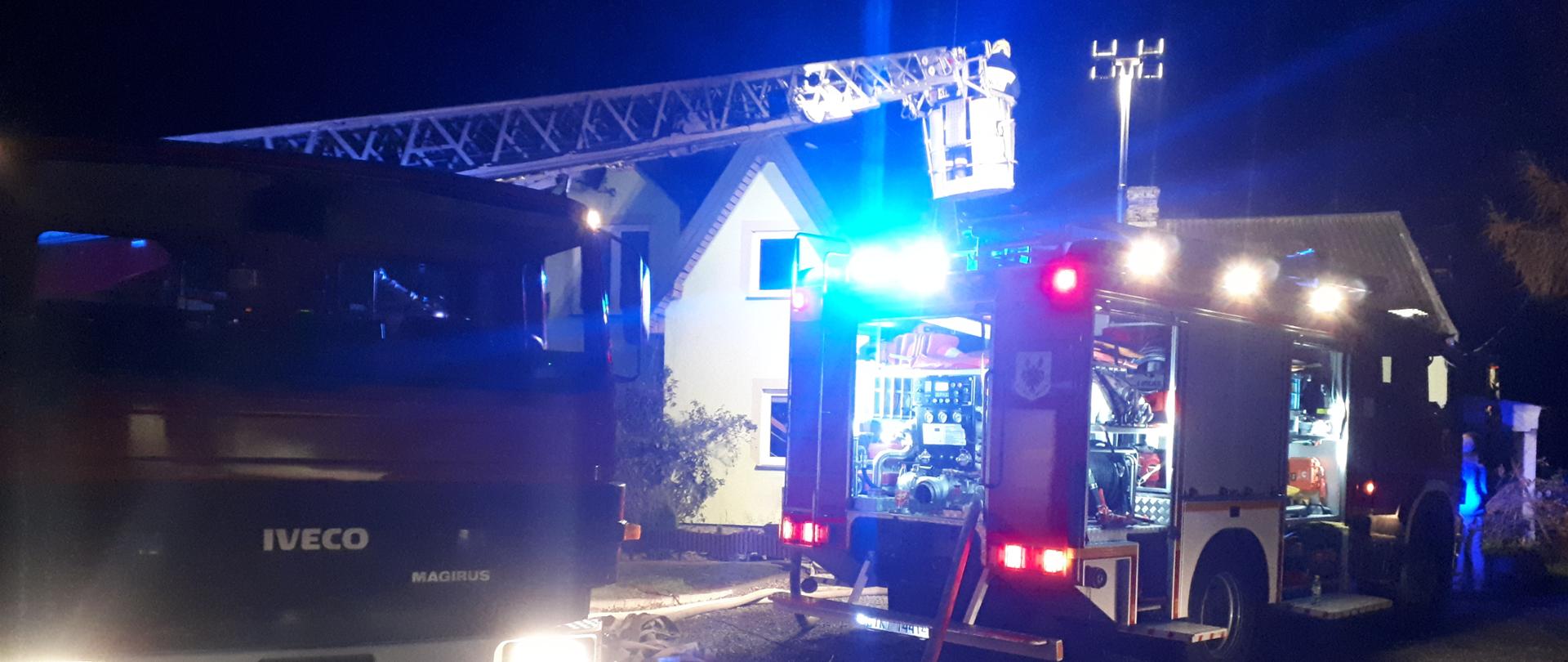 Zdjęcie przedstawia samochody gaśnicze oraz rozstawioną drabinę mechaniczną na miejscu zdarzenia. Przed samochodami znajdują się rozwinięte węże gaśnicze. Nie widać ognia ani dymu. Na zewnątrz jest ciemno.