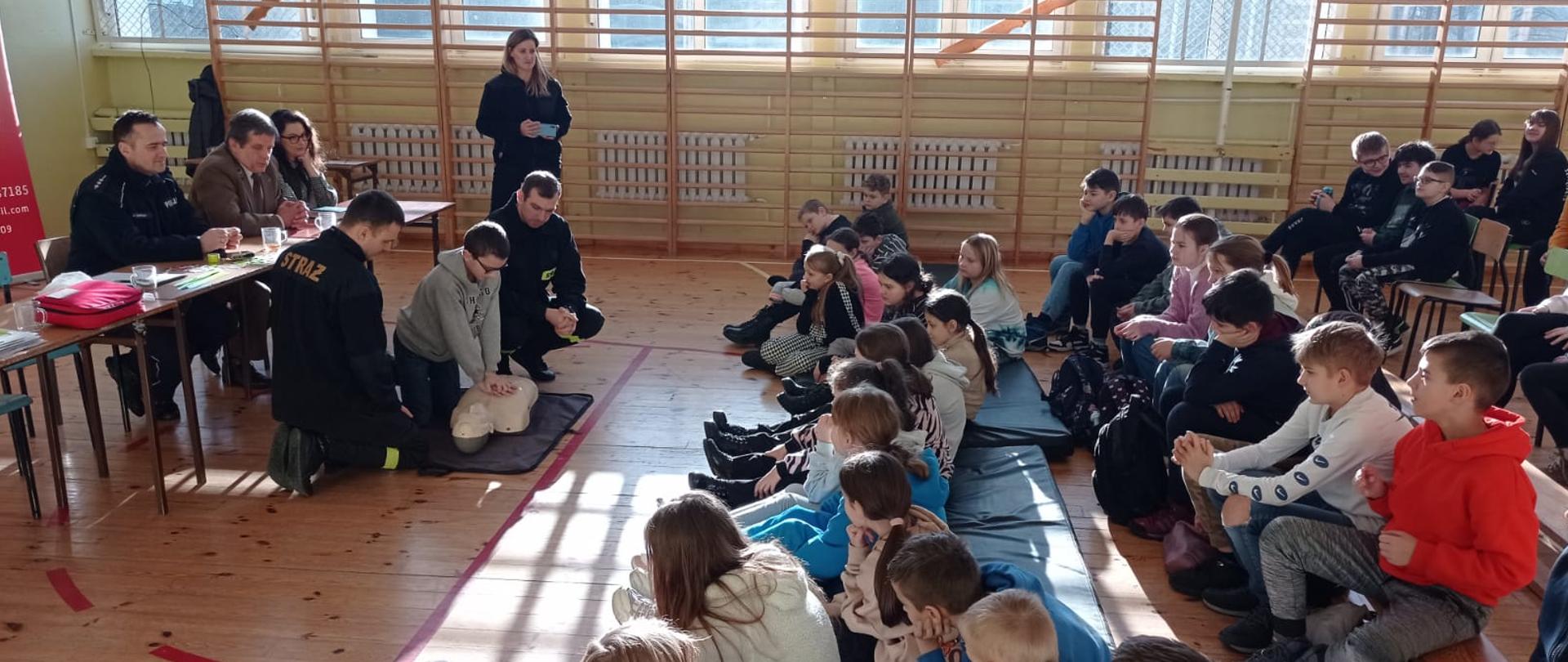 Zdjęcie przedstawia grupę dzieci w sali gimnastycznej podczas pogadanki na temat bezpieczeństwa ze strażakami, Policjantami oraz leśnikami.