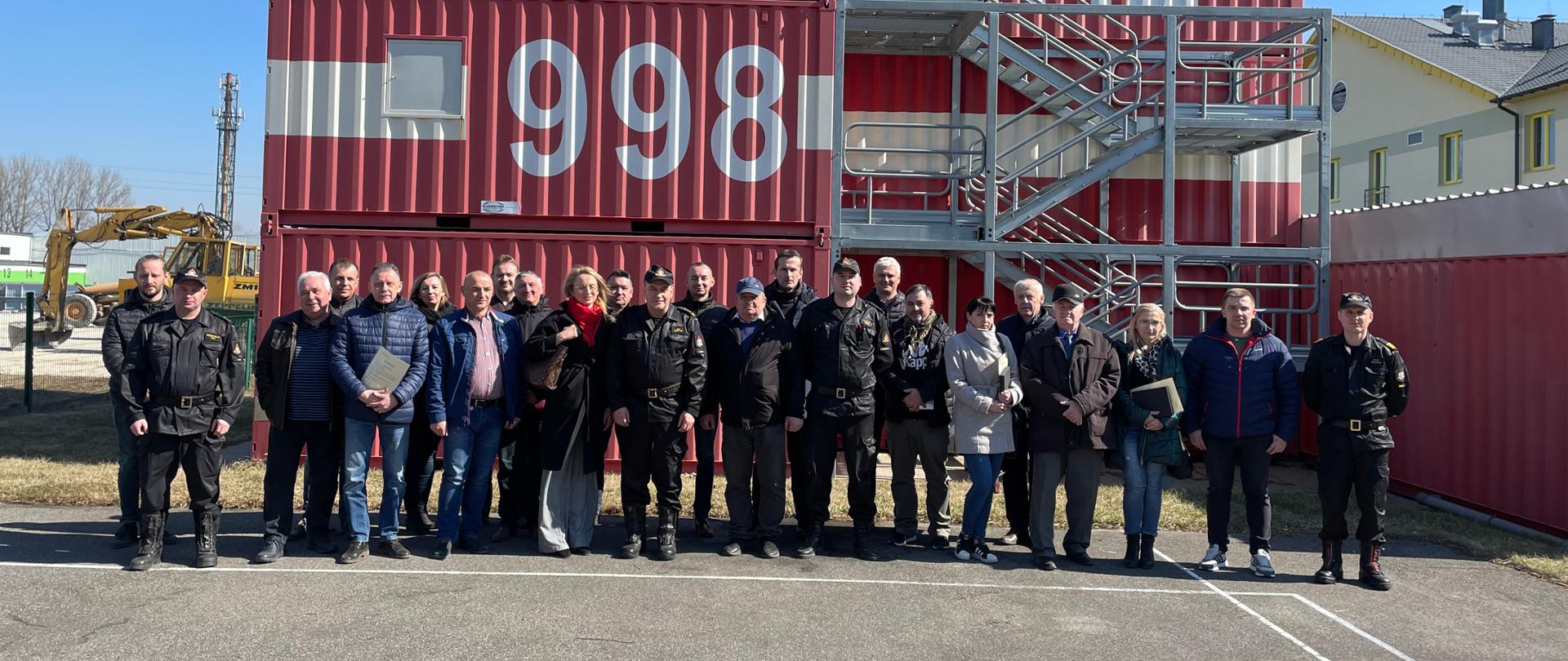 Zdjęcie przedstawia pamiątkową fotografię uczestników szkolenia wraz z kadrą Ośrodka Szkolenia KW PSP w Kielcach. Uczestnicy są zadowoleni z pomyślnego zdania egzaminu. Kadra ośrodka szkolenia ubrana jest w mundury dowódczo-sztabowe. 