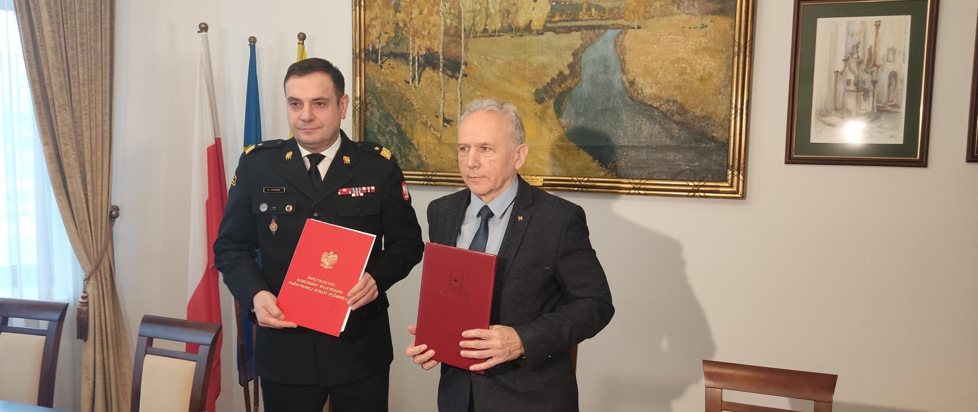 Na zdjęciu widzimy dwóch mężczyzn pozujących do zdjęcia z teczkami koloru czerwonego. Po lewej stronie strażak w mundurze wyjściowym w stopniu nadbrygadiera PSP, po prawej rektor politechniki w garniturze. Za ich plecami obraz i flagi.