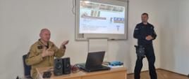 Strażak w mundurze musztardowym siedzi za biurkiem na którym są rozłożone głośniki laptop na ekranie z rzutnika wyświetlana jest prezentacja obok stoi funkcjonariusz Policji.