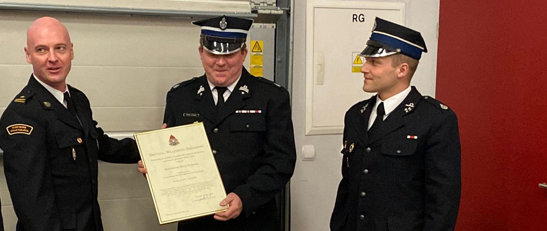 Zdjęcie przedstawia Komendanta Powiatowego PSP w Jędrzejowie mł. bryg. Mariusza Stolarczyka, który wręcza przedstawicielom OSP Sędziszów decyzję o włączeniu do krajowego systemu ratowniczo-gaśniczego. Wszyscy strażacy w umundurowaniu galowym znajdują się na garażu jednostki OSP Sędziszów.