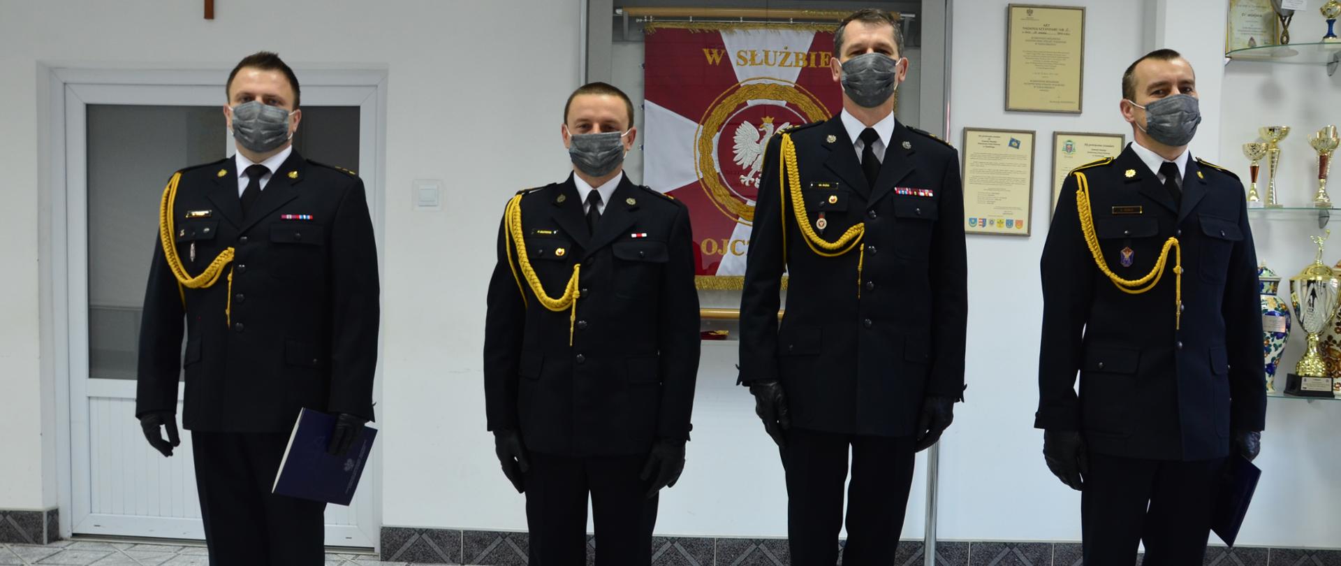 Zdjęcie przedstawia czterech strażaków w mundurach galowych, stojących w szeregu w świetlicy Komendy Miejskiej Państwowej Straży Pożarnej w Tarnobrzegu. 