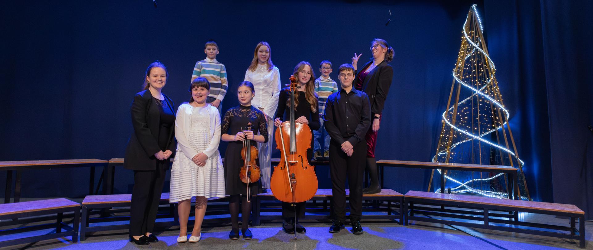 Na pamiątkowym zdjęciu z koncertu w Orzeszu widać grupę osób stojących na scenie. Przednia część grupy składa się z 5 młodych osób ubranych elegancko — niektórzy trzymają instrumenty muzyczne, takie jak skrzypce i wiolonczela.
W tle, na podwyższeniu, stoją 4 inne osoby, również odświętnie ubrane. Scena jest oświetlona niebieskim światłem, a po prawej stronie znajduje się dekoracja przypominająca choinkę z lampek, co nadaje zdjęciu świąteczny charakter. 