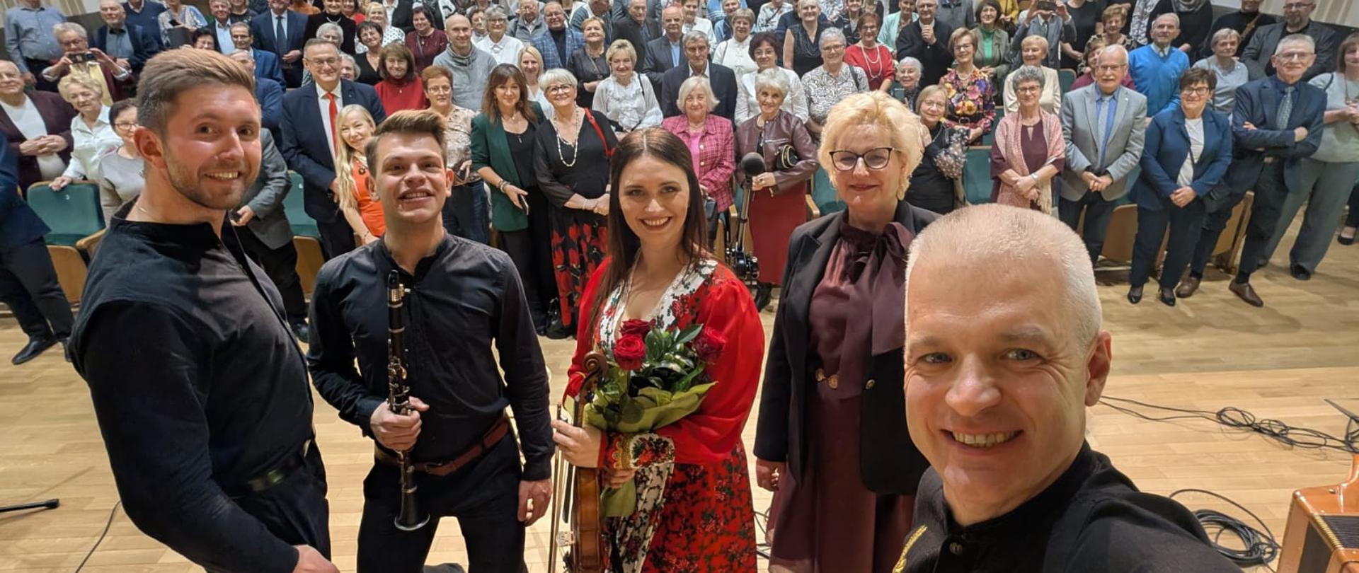 Na zdjęciu artyści, którzy wystąpili podczas koncertu. Trzech mężczyzn ubranych na czarno oraz kobieta w długiej czerwonej sukni z szerokim uśmiechem, która trzyma czerwone róże. Obok niej kobieta blondynka w brązowej sukience i czarnej marynarce. W tle na widowni licznie zgromadzona publiczność, która uczestniczyła w I Powiatowych Obchodach Dnia Seniora.