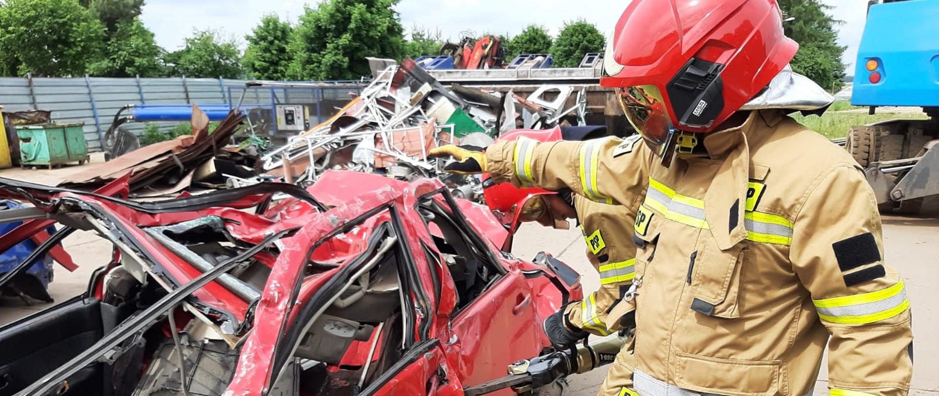 Na zdjęciu widać wrak samochodu a przy nim 2 strażaków wycinających drzwi