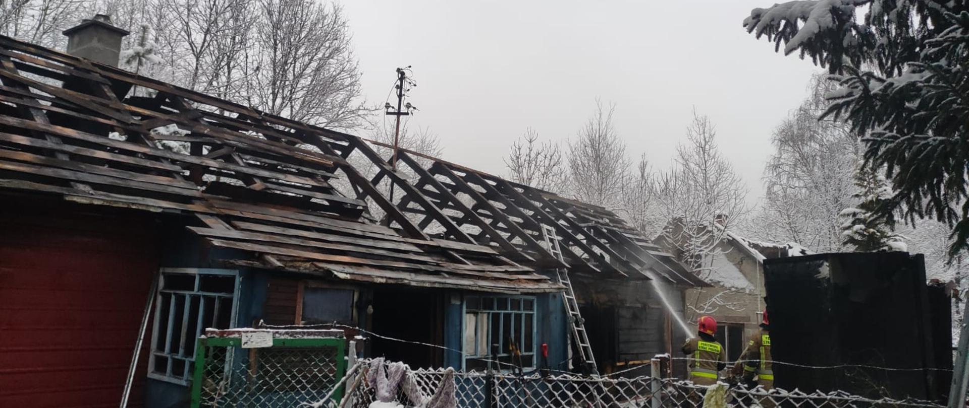 Zdjęcie przedstawia zgliszcza domu drewnianego, dogaszanego przez strażaków. Widoczna zwęglona więźba dachowa. 