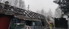 Zdjęcie przedstawia zgliszcza domu drewnianego, dogaszanego przez strażaków. Widoczna zwęglona więźba dachowa. 