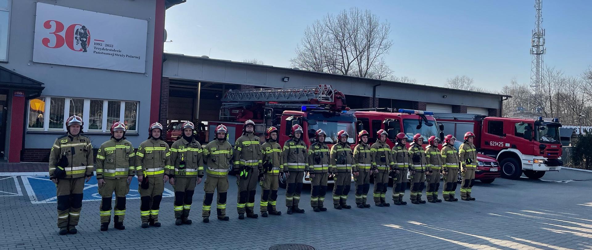 Zdjęcie przedstawia strażaków stojących w szeregu przed budynkiem Komendy Miejskiej Państwowej Straży Pożarnej w Świętochłowicach, w tle samochody pożarnicze.