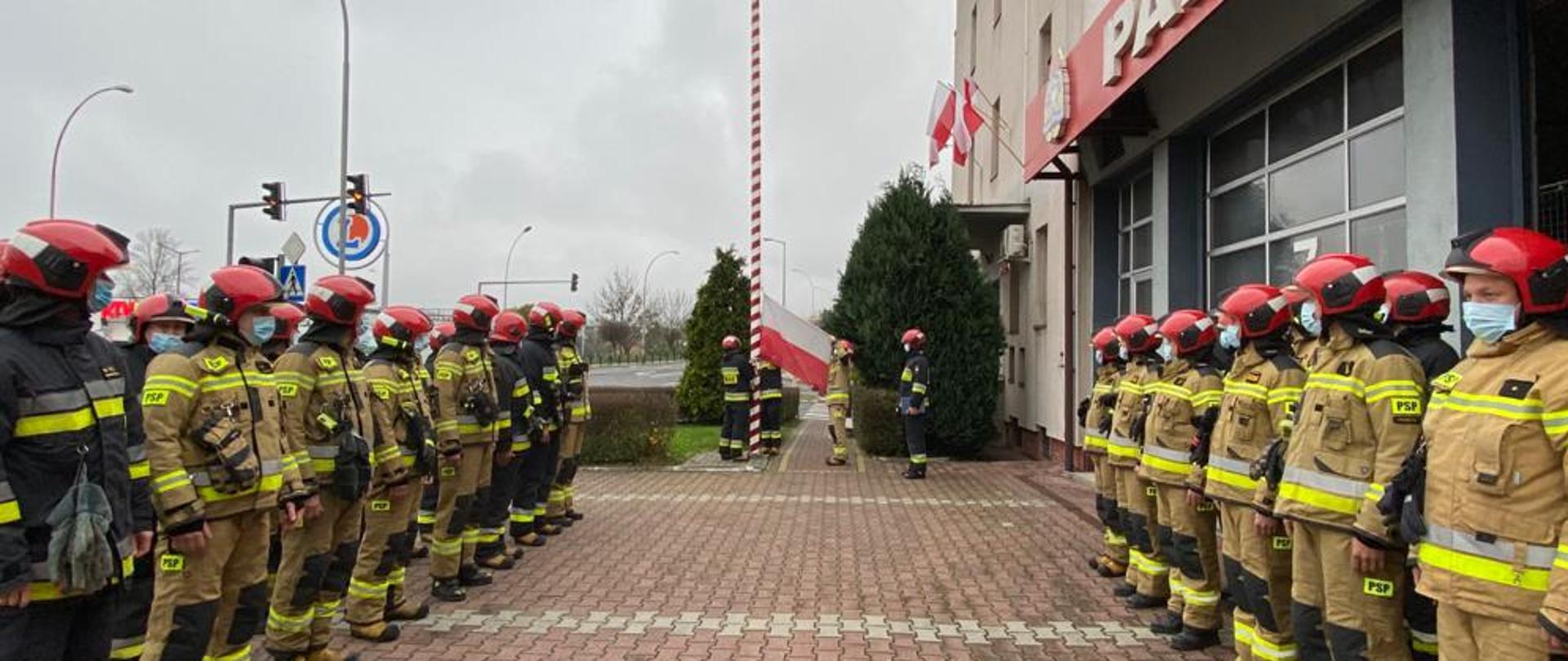 Przemyscy strażacy uczcili Narodowe Święto Niepodległości. Zbiórka w mundurach specjalnych na placu zewnętrznym, w tle podnoszenie Flagi Państwowej. 