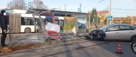 Na zdjęciu przedstawiono miejsce wypadku drogowego, gdzie samochód osobowy uderzył w wiatę przystanku autobusowego. Konstrukcja wiaty została poważnie uszkodzona, a wokół widoczne są rozbite elementy metalowe oraz szkło. W tle znajduje się autobus oraz osoba w ciemnym ubraniu obserwująca miejsce zdarzenia. Na jezdni ustawiony jest pomarańczowy słupek ostrzegawczy, a w pobliżu przystanku przebiegają tory tramwajowe.
