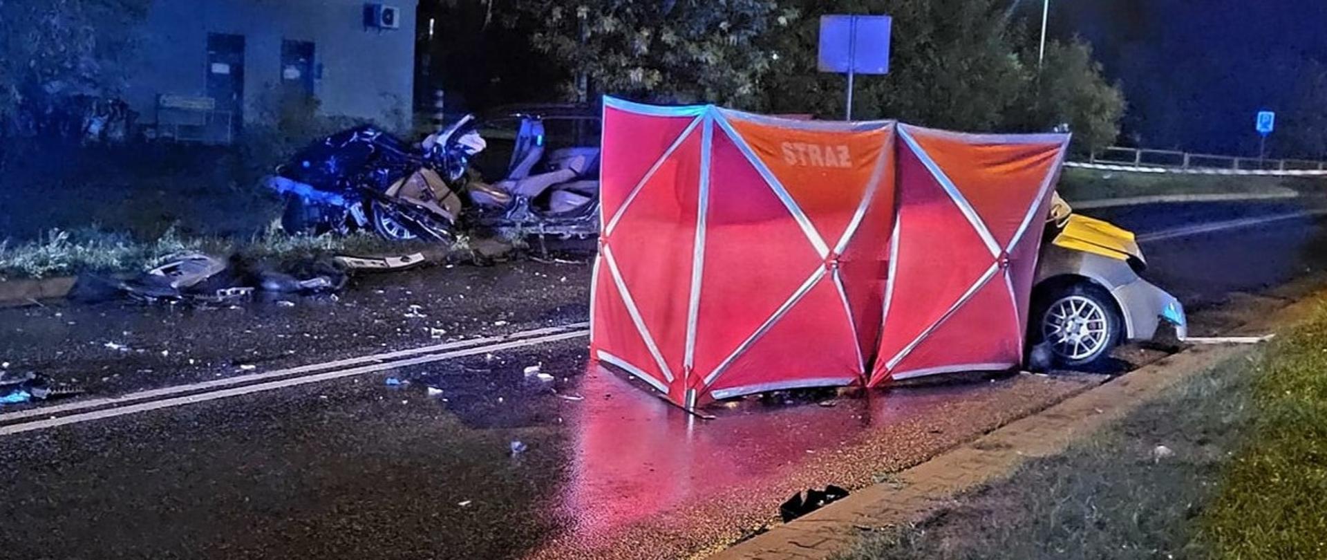 Zdjęcie przedstawia zderzenie dwóch samochodów osobowych. Jeden pojazd zasłonięty jest parawanem z napisem straż.